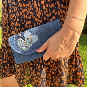 Peut inclure: Une pochette en jean avec un appliqué en forme de cœur. L'appliqué présente un cœur bleu clair avec le mot "June" brodé dessus, et est orné de clous dorés. La pochette est tenue par une personne.