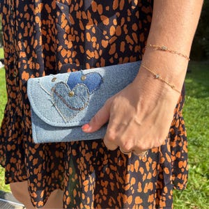Peut inclure: Une pochette en jean bleu clair avec un motif de cœur composé de pièces de jean et de clous en laiton. La pochette est tenue devant une robe avec un fond noir et des taches orange. Un bracelet en or est également visible.