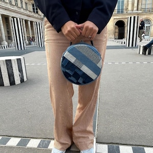 Peut inclure: Un sac à main rond en denim avec un motif patchwork dans différentes nuances de bleu. Le sac a une poignée noire. La personne porte un pantalon en velours côtelé beige. L'arrière-plan présente un bâtiment avec des colonnes rayées noires et blanches.
