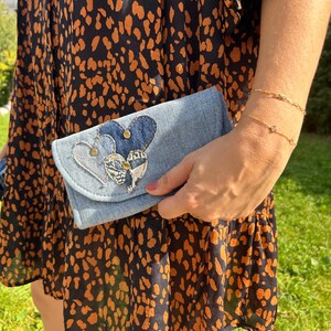 Peut inclure: Une pochette en jean bleu clair avec trois appliqués en forme de cœur. Les cœurs sont en différentes nuances de denim bleu et ornés de clous dorés. La pochette est tenue contre une robe à motifs tachetés noirs et orange.