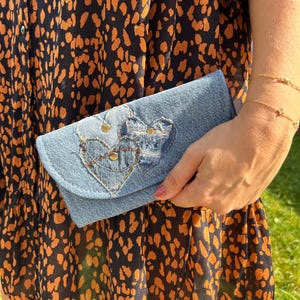 Peut inclure: Une pochette portefeuille en jean bleu clair ornée de deux appliqués en forme de cœur aux bords effilochés, de coutures décoratives et de petits clous métalliques. Le portefeuille possède un rabat et est fait de tissu denim en coton bleu.