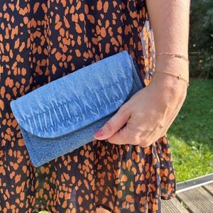 Peut inclure: Une pochette en jean bleu clair avec un rabat froncé. La pochette est tenue contre une robe à motifs avec un fond noir et des taches orange. Un bracelet doré est visible au poignet.