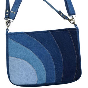 May include: A denim crossbody bag with a patchwork design in shades of blue. The bag features a curved pattern and a long, adjustable strap with metal hardware. The bag is approximately 10 inches wide and 8 inches tall.