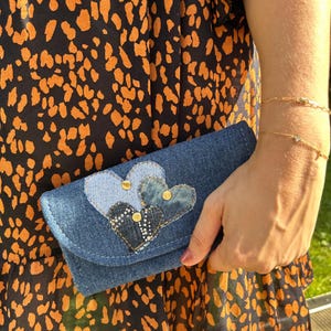 Peut inclure: Pochette en jean avec trois appliqués en forme de cœur de différentes nuances de bleu. Le plus grand cœur est orné de petits clous argentés et de rivets dorés. La pochette est tenue contre une robe à motifs orange et noir.