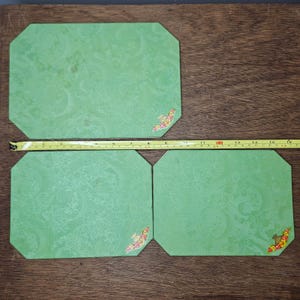 Puede incluir: Tres manteles individuales verdes con un estampado floral y detalles decorativos en las esquinas. Cada mantel individual mide aproximadamente 38 cm de largo. Los manteles individuales tienen forma octogonal y están colocados sobre una superficie de madera.