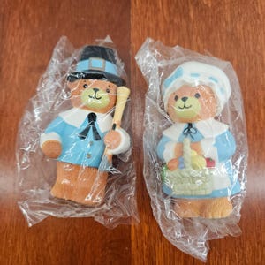 May include: Two ceramic bear figurines, one male and one female, dressed in Pilgrim attire. The male bear wears a black hat and holds a staff. The female bear wears a bonnet and holds a basket. Both are in plastic packaging.
