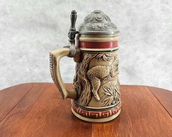 May include: A beige ceramic beer stein with a detailed relief of a leopard resting in a tree. The stein has a metal hinged lid and handle. The design includes a red and green border.