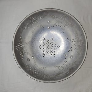 Puede incluir: Un colador de metal plateado con un patrón de agujeros en forma de estrella en el centro y alrededor de los bordes. El colador es redondo con un borde ligeramente ensanchado y está diseñado para escurrir líquidos de los alimentos.