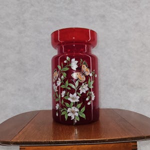 Op de afbeelding: Een robijnrode glazen vaas met een cilindrische body en een uitlopende bovenkant. De vaas is versierd met witte bloemen, groene bladeren en oranje en bruine vlinders. De vaas staat op een houten tafel.