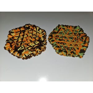 May include: Two handmade, round coasters. One coaster features a pattern of orange, yellow, and brown yarn. The other coaster has a similar design, but with green accents. The coasters are made of a textured, woven material.