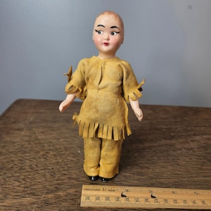 Può includere: Una bambola vintage con il viso dipinto, che indossa un abito in pelle marrone con frange e accenti decorativi. L'abito della bambola comprende una tunica e pantaloni. La bambola è in piedi su una superficie di legno. Un righello è in primo piano.