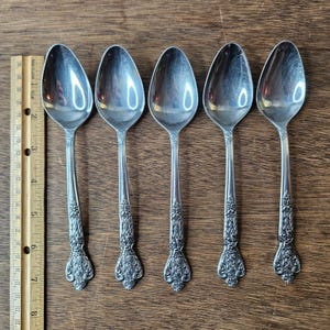 Puede incluir: Cinco cucharas plateadas con detalles florales ornamentados en los mangos. Las cucharas están dispuestas en fila sobre una superficie de madera. Una regla de madera se coloca junto a las cucharas.