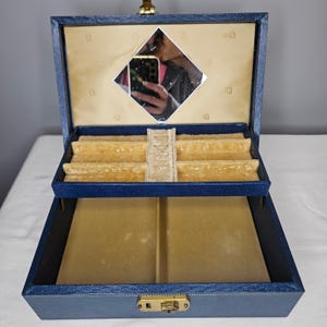 Puede incluir: Un joyero azul marino con exterior texturizado y cierre de metal dorado. El interior cuenta con un forro de terciopelo beige, una bandeja extraíble con múltiples compartimentos para anillos y objetos pequeños, y un espejo en forma de rombo.
