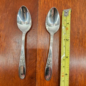 Peut inclure: Deux cuillères en argent avec des détails floraux ornementaux sur les manches. Les cuillères mesurent environ 15 cm de long. Un ruban à mesurer jaune est à côté des cuillères.