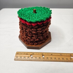 May include: A cylindrical crocheted object with a brown textured body, a red middle layer, and a green top, resting on a hexagonal wooden base. A wooden ruler is placed in front, showing the object is approximately 3.5 inches in diameter.