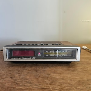 May include: A vintage Realistic Chronomatic-233 digital alarm clock. The clock has a red LED display, a silver face, and a wood-grain top. It features AM/FM radio with a frequency dial and a battery indicator.