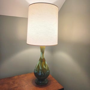 Puede incluir: Una lámpara de mesa con una pantalla cilíndrica de tela color blanco roto y una base de cerámica en forma de lágrima. La base presenta un esmalte brillante de varios tonos con vetas mezcladas de verde oscuro, oliva y crema en un patrón.
