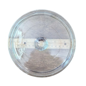 Op de afbeelding: Een doorzichtige glazen deksel met een ronde vorm en een kleine, ronde handgreep in het midden. Een houten liniaal met inch-markeringen ligt eronder. De deksel is ontworpen voor potten of pannen.