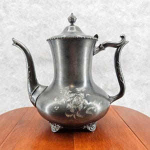 May include: Antique silver-colored teapot with a curved spout and handle. The teapot features a floral design on the body and sits on three decorative feet. The lid is topped with a small finial.