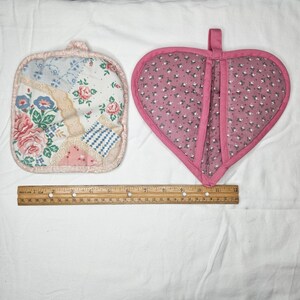 May include: Two quilted pot holders. One is square with floral and patchwork patterns, the other is heart-shaped with a pink floral print. A wooden ruler is placed below the pot holders. The square pot holder measures approximately 7 inches.