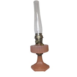 May include: A vintage-style oil lamp featuring a pale pink, ribbed glass base and font. The lamp includes a metallic burner assembly with a circular adjustment knob and a tall, clear, cylindrical glass chimney extending from the top of the burner.