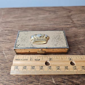 Könnte beinhalten: Eine rechteckige, goldfarbene Metallbox mit einer dekorativen goldenen Krone auf dem Deckel. Die Box ist etwa 5 cm lang. Ein Holzlineal mit "MADE IN JAPAN" liegt neben der Box.