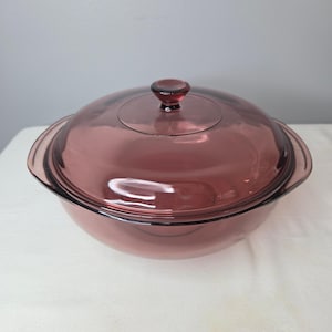 May include: A round, translucent plum-colored glass casserole dish with a matching lid. The dish features integrated side handles and a central knob on the lid. The glass has a smooth, polished finish, allowing light to pass through the deep purple hue.