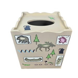 May include: Beige wooden tissue box with a square shape and a scalloped top. The box is decorated with forest-themed illustrations, including a bear, moose, canoe, and trees. The illustrations are in shades of green, brown, and blue.