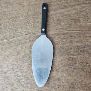 May include: A stainless steel cake server with a black handle. The server has a wide, flat blade and a black handle with a small white dot near the top and bottom. The server is designed for cutting and serving cakes and pies.