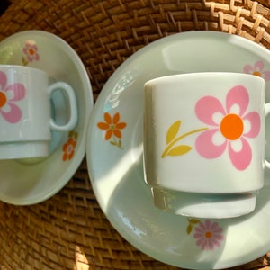 Könnte beinhalten: Zwei weiße Keramik-Teetassen mit rosa und orangefarbenen Blumendesigns auf einem weißen Unterteller. Die Teetassen haben ein schlichtes, elegantes Design.