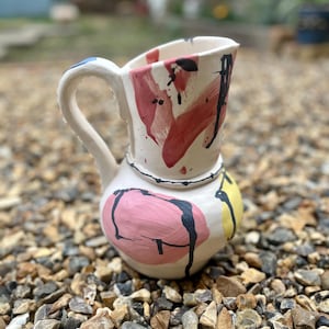 Peut inclure: Pichet en céramique fait à la main, avec une base blanche et des coups de pinceau abstraits rouges, roses, noirs et jaunes. La poignée est blanche avec un accent bleu. Le pichet est posé sur un lit de petits cailloux.