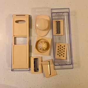 May include: A set of beige plastic kitchen tools for slicing, grating, and julienning vegetables. The set includes a base with a clear plastic container, a grater, a julienne slicer, a mandoline, and a slicer with a round blade. The base has the words "Mini Wonder" printed on it.