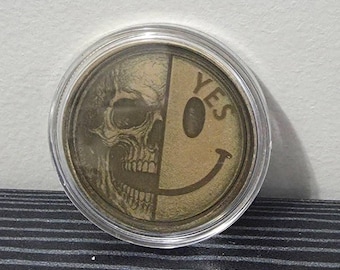 May include: A circular gold-toned metal coin encased in a clear plastic protective capsule. The coin features a split design with a detailed human skull on the left side and a classic smiley face with the word YES printed on the right side.