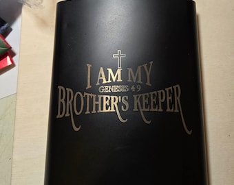 May include: A matte black metal flask with a silver-toned screw cap. The front features an engraved design with a cross symbol above the text "I AM MY BROTHER'S KEEPER" and "GENESIS 4 9" in a light-colored font that contrasts with the dark surface.