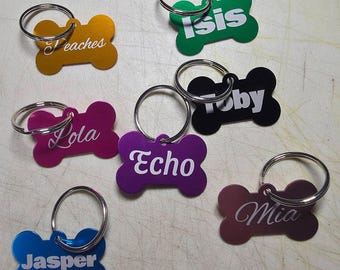 May include: A collection of seven bone-shaped metal pet ID tags in various colors including gold, green, black, magenta, purple, blue, and maroon. Each tag features a silver split ring and white engraved names: Peaches, Isis, Toby, Lola, Echo, Mia, and Jasper.
