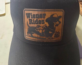 May include: A black baseball cap featuring a rectangular brown leather patch on the front. The patch displays a graphic of a dachshund wearing a cowboy hat and saddle, with the text "Wiener Rides" and "Only 25¢" printed in black.