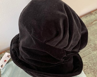 May include: A black velvet bucket hat with a gathered crown. The hat is sitting on a light blue fabric surface.