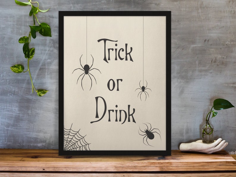 K&ouml;nnte beinhalten: Ein gerahmter Druck mit dem Text "Trick or Drink" in einer dekorativen Schriftart. Drei schwarze Spinnen-Silhouetten h&auml;ngen oben am Druck. Ein Spinnennetz-Design befindet sich unten am Druck.