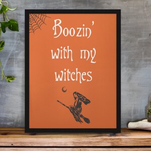 K&ouml;nnte beinhalten: Orangener Hintergrund mit schwarzem Rahmen und einer wei&szlig;en Silhouette einer Hexe, die auf einem Besen fliegt. Der Text "Boozin' with my witches" ist in wei&szlig;er Kursivschrift geschrieben.