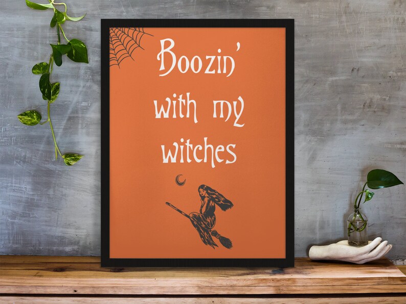 K&ouml;nnte beinhalten: Orangener Hintergrund mit schwarzem Rahmen und einer wei&szlig;en Silhouette einer Hexe, die auf einem Besen fliegt. Der Text "Boozin' with my witches" ist in wei&szlig;er Kursivschrift geschrieben.