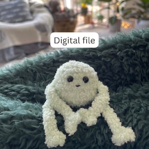 Op de afbeelding: Een lichtgroen gehaakt pluche speelgoed met grote zwarte ogen en een kleine glimlach. Het speelgoed heeft lange, pluche armen en benen en zit op een groen, pluche oppervlak. Digitaal bestand.