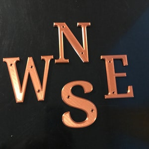 May include: Four copper-colored metal letters spelling out the word "W.S.N.E."  The letters are arranged in a circle, with the "W" at the top, the "S" at the bottom, the "N" on the right, and the "E" on the left.  Each letter has two small holes in it.