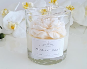 May include: A white soy wax candle in a clear glass jar with a white flower on top. The candle is labelled "Aromatic Candle" and "Handmade".