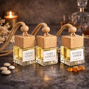 Op de afbeelding: Drie glazen flessen met houten doppen en labels met de tekst "FRESH LINEN", "VERY VANLLA" en "SANDAL AMBER". Elke fles heeft een beige koord en een houten kraal. De flessen staan op een donkere ondergrond met kleine stenen en barnsteen.