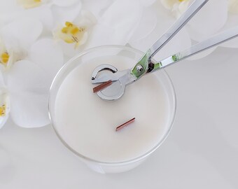 May include: A close-up of a white candle in a glass jar with a silver candle wick trimmer. The trimmer is holding a copper wick.