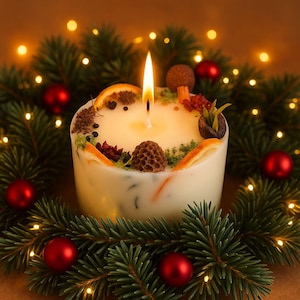 Op de afbeelding: Een brandende witte pilaar kaars versierd met gedroogde sinaasappelschijfjes, kruiden en een kleine dennenappel. De kaars is omringd door een krans van groenblijvende takken en rode ballen, met kleine lichtjes op de achtergrond.
