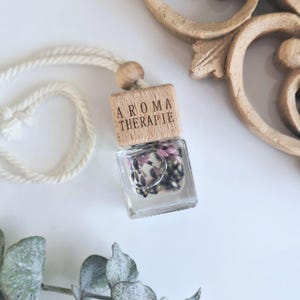 Könnte beinhalten: Ein klarer Glas-Aromatherapie-Diffusor mit getrockneten Blumen im Inneren, gekrönt von einem Holzdeckel mit der Aufschrift "AROMA THERAPIE". Der Diffusor hängt an einem weißen Seil. Die Gesamtästhetik ist natürlich und beruhigend.