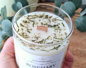 May include: A clear glass jar candle with a white soy wax candle inside. The candle has a wooden wick and is decorated with dried herbs. The label on the jar reads "Soy Wax Scented Candle Rosemary Sage by Gather Home".