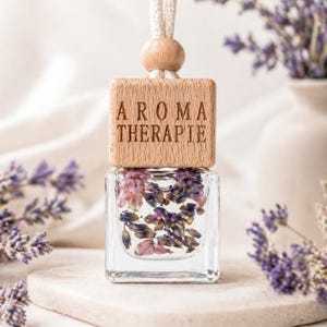 Könnte beinhalten: Ein klarer Glas-Aromatherapie-Diffusor mit getrockneten Lavendelblüten im Inneren. Der Holzdeckel ist mit "AROMA THERAPIE" graviert. Eine Holzkugel und eine weiße Schnur sind zum Aufhängen befestigt.