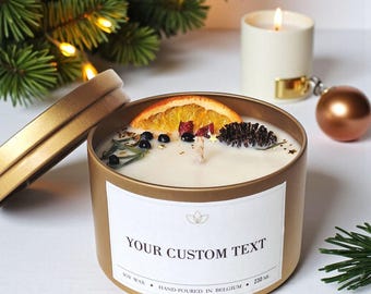 May include: A gold-colored candle tin with a lit candle inside, decorated with a dried orange slice, berries, and a pine cone. The label reads "YOUR CUSTOM TEXT". A second lit candle and Christmas ornaments are in the background.